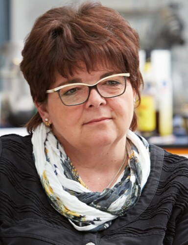 Mittleren Alters sitzende Frau mit kurz geschnittener brünetter Frisur und Brille, die nachdenklich in die Ferne blickt und einen gemusterten Schal über dunkler Kleidung trägt.