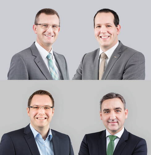 Vier männliche Geschäftsleute in zwei Reihen posieren im Anzug vor neutralgrauem Hintergrund und lächeln professionell in die Kamera