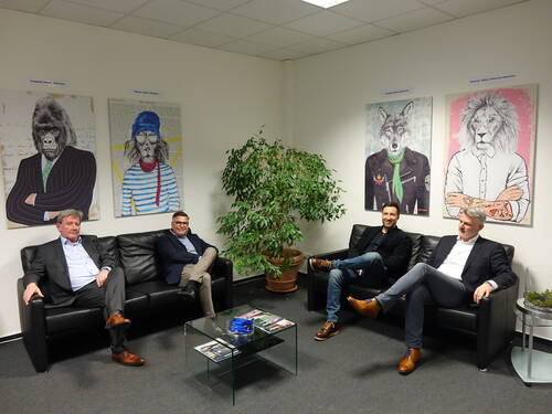 Vier Männer in Businesskleidung sitzen entspannt auf zwei schwarzen Ledersofas in einem modern eingerichteten Büro, flankiert von großformatigen, humorvollen Tierporträts in Menschenkleidung an der Wand.
