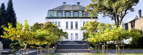 Elegante weiße Villa mit grünem Mansardendach, von symmetrischen Treppen und herbstlich gefärbten Bäumen im gepflegten Vordergarten eingerahmt