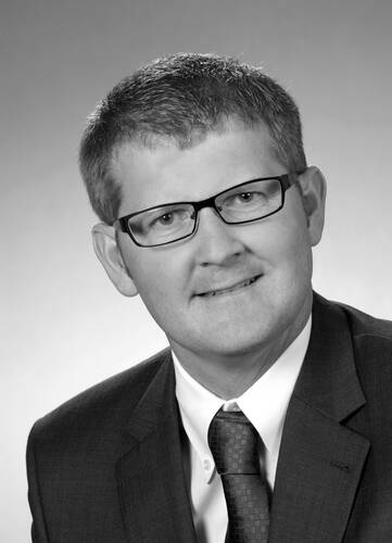 Mittelalterlicher Mann mit kurzem Haar und Brille lächelt leicht in einem formellen Schwarz-Weiß-Bewerbungsfoto im Anzug mit Krawatte.