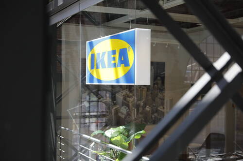 Leuchtendes IKEA-Schild hängt vor einer Glaswand in einem modernen Innenraum mit Pflanzen und Stadtposter im Hintergrund