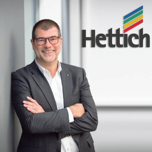 Geschäftsmann im Anzug steht lächelnd mit verschränkten Armen vor einer Wand mit dem Hettich-Unternehmenslogo.