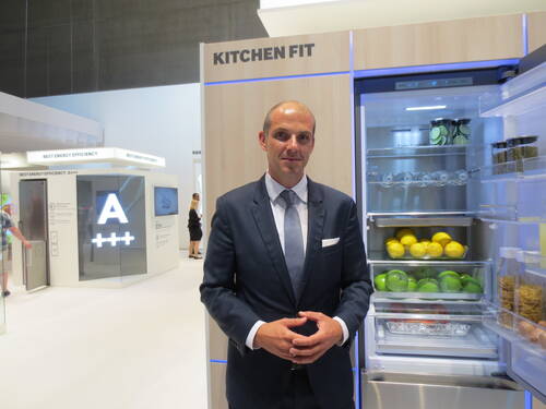 Geschäftsmann im Anzug präsentiert auf einer Messe einen geöffneten, energieeffizienten Kühlschrank mit ordentlich sortiertem Obst und Lebensmitteln