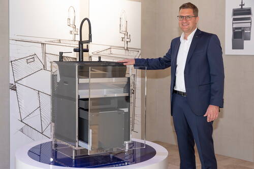 Geschäftsmann im Anzug präsentiert in einer Ausstellung ein durchsichtiges Küchenunterschrank-Modell mit integriertem Spülbecken und schwarzer Armatur vor technischen Zeichnungen im Hintergrund.