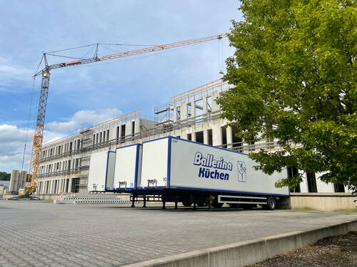 Große Baustelle mit mehrstöckigem Rohbau, gelbem Turmdrehkran und LKW-Anhängern der Marke „Ballerina Küchen“ auf einem gepflasterten Hof neben einem grünen Baum