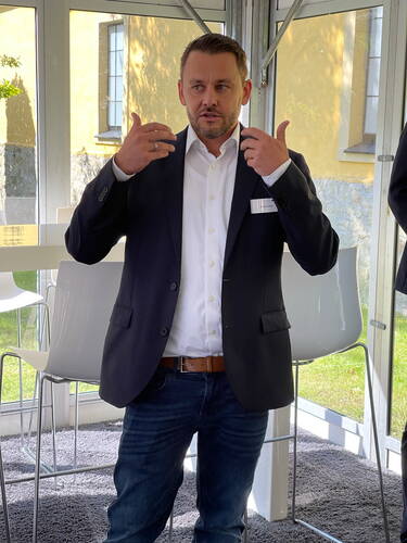 Geschäftsmann im Anzug spricht gestikulierend in einem hellen, modernen Seminarraum mit Glaswänden und Barhockern.