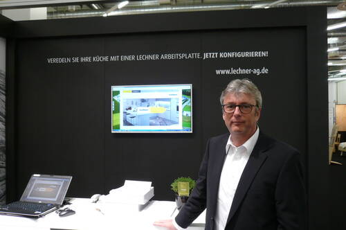 Mann im Anzug steht an einem modernen Messe­stand von Lechner mit Laptop, Monitor und Küchenarbeitsplatten-Konfigurator im Hintergrund