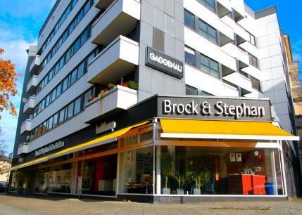 Moderne mehrstöckige Wohn- und Geschäftshausfassade mit dem Küchenstudio „Brock & Stephan“ im Erdgeschoss und gelber Markise an einer sonnigen Straßenecke