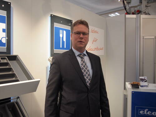Geschäftsmann im Anzug steht an einem Messestand mit Küchenausstattung und blauem Bestecksymbol im Hintergrund