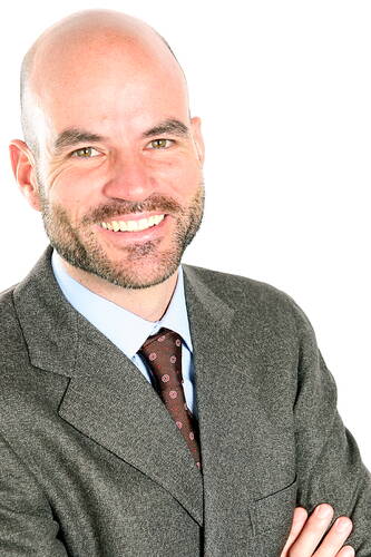 Lächelnder, glatzköpfiger Mann im Anzug mit gekreuzten Armen vor weißem Hintergrund
