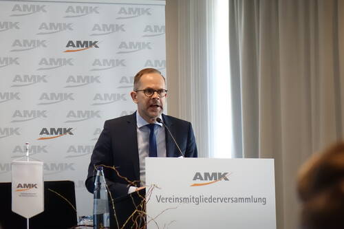 Mann im Anzug spricht am Rednerpult der AMK während einer Vereinsmitgliederversammlung in einem Konferenzraum.