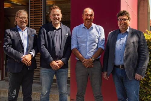 Vier lächelnde Männer in Business-Kleidung stehen nebeneinander im Sonnenschein vor einem modernen Gebäude mit roter Fassade und Holzelementen.