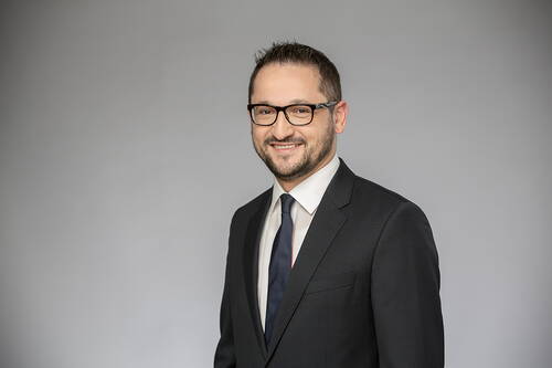 Lächelnder Mann mit Brille im dunklen Business-Anzug und Krawatte vor neutral grauem Hintergrund