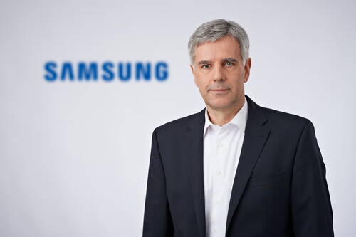 Geschäftsmann im dunklen Anzug mit weißem Hemd steht vor unscharfem Samsung-Logo auf hellem Hintergrund