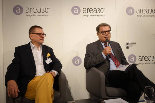 Zwei Geschäftsleute in Anzügen sitzen auf einer Podiumsdiskussion bei der Messe area30, während der rechte Mann mit Mikrofon spricht und Unterlagen hält.
