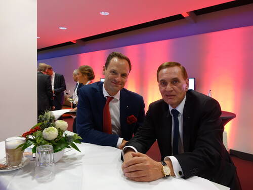 Zwei elegant gekleidete Männer in Anzügen sitzen an einem weiß gedeckten Tisch bei einer Abendveranstaltung mit rot-violetter Ambientebeleuchtung und Blumenarrangement im Vordergrund.