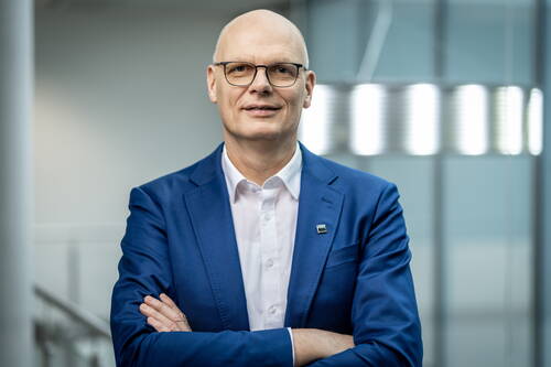 Glatzköpfiger Mann mit Brille im blauen Anzug und verschränkten Armen steht selbstbewusst in einem modernen Bürogebäude