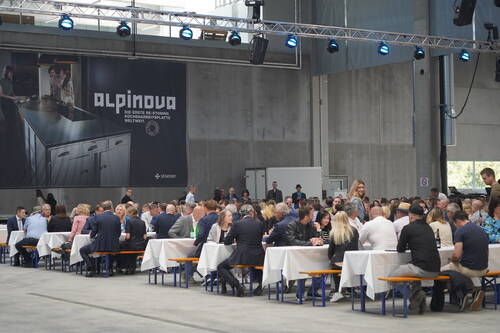Große Firmenveranstaltung in einer Industriehalle, bei der zahlreiche Gäste an langen, weiß gedeckten Tischen sitzen und vor einer Wand mit großem „alpinova“-Werbebanner speisen und diskutieren.