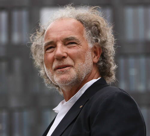 Älterer Mann mit grauen Locken und Bart blickt im dunklen Sakko vor einem modernen Glasgebäude in die Kamera