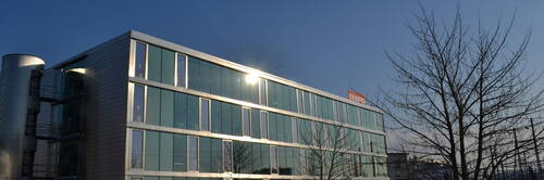 Modernes Bürogebäude mit gläserner Fassade in der Abendsonne, flankiert von kahlen Bäumen im Vordergrund