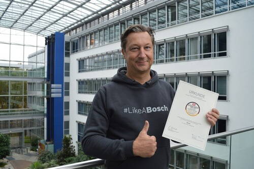 Mann im #LikeABosch‑Pullover steht in moderner Büroatrium-Architektur, hält stolz eine Urkunde in der einen Hand und zeigt mit der anderen den Daumen nach oben