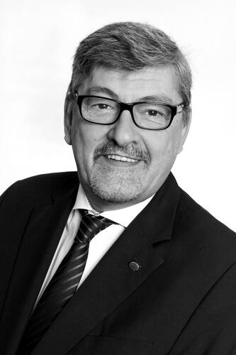 Älterer Mann mit Brille und Schnurrbart lächelt im schwarzen Anzug mit gestreifter Krawatte in einem formellen Porträt in Schwarz-Weiß