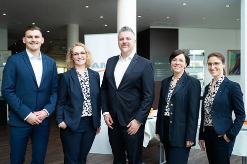 Fünf Business‑Personen in dunklen Anzügen stehen lächelnd nebeneinander in einem modernen Konferenzraum vor einem Firmenbanner.