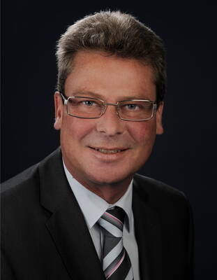 Mittelälterer Mann mit Brille im dunklen Anzug und gestreifter Krawatte lächelt vor dunklem Hintergrund in die Kamera
