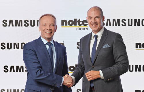 Zwei Geschäftsleute im Anzug lächeln und schütteln sich vor den Logos von Samsung und Nolte Küchen die Hand, während sie eine Partnerschaft besiegeln.