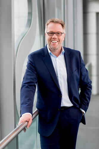 Lächelnder Mann im dunkelblauen Anzug lehnt sich in einem modernen Bürogebäude lässig an ein Geländer und schaut direkt in die Kamera.