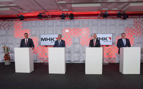 Vier Manager im Anzug stehen hinter Rednerpulten vor einer beleuchteten Bühnenwand mit MHK-Group-Logos während einer Unternehmensveranstaltung
