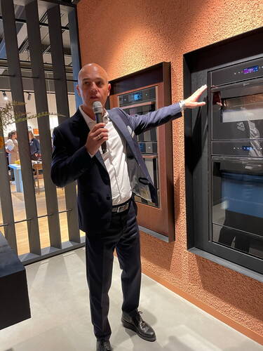 Mann im Anzug erklärt mit Mikrofon in der Hand ein modernes, in die Wand eingebautes Küchengerät in einer Ausstellungsumgebung