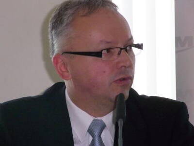 Mittelalterlicher Mann im Anzug und mit Brille spricht konzentriert in ein Mikrofon während einer formellen Veranstaltung