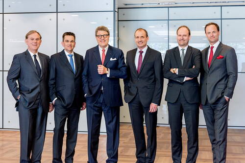 Sechs Geschäftsleute in Anzügen stehen nebeneinander in einem modernen Konferenzraum und lächeln in die Kamera