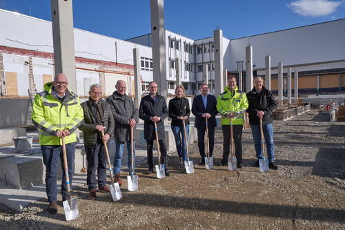 Gruppe von acht Personen mit Spaten beim symbolischen ersten Spatenstich auf einer Baustelle vor einem modernen Industrie- oder Bürogebäude bei sonnigem Wetter