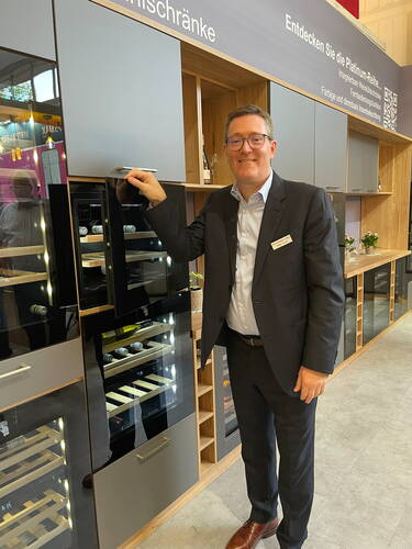 Geschäftsmann im Anzug präsentiert eine moderne Einbauküche mit integrierten Weinkühlschränken auf einer Fachmesse