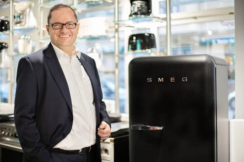 Geschäftsmann im Anzug lächelt neben einem schwarzen SMEG-Kühlschrank in einem modernen Haushaltsgeräte-Showroom
