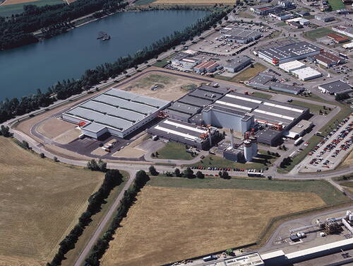 Großes Industrie- und Logistikgelände mit Hallen, Silos und Parkplätzen an einem Fluss, umgeben von Feldern und weiteren Gewerbebauten