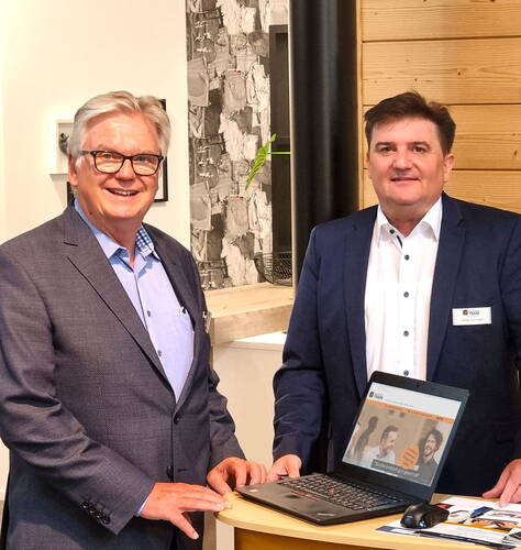 Zwei geschäftlich gekleidete Männer stehen lächelnd an einem runden Stehtisch und präsentieren gemeinsam einen geöffneten Laptop in einem modernen Büro- oder Messeraum.