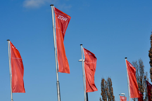 Mehrere rote ZOW-Flaggen wehen an hohen Masten vor einem klaren blauen Himmel neben schlanken Bäumen.