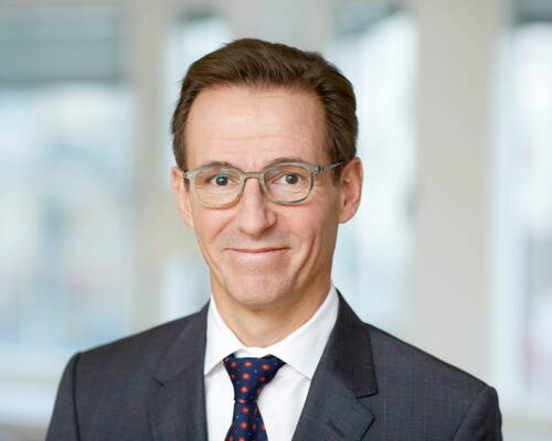 Mittelälterer Mann mit Brille und dunklem Anzug lächelt dezent in einer hellen, modernen Büro Umgebung in die Kamera