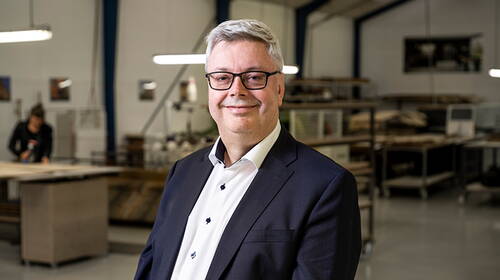 Älterer Mann im Anzug und mit Brille lächelt in einer Werkhalle, während im Hintergrund eine Person an einem Arbeitstisch beschäftigt ist.