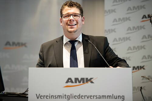 Lächelnder Mann im Anzug spricht am Rednerpult mit AMK-Logo während einer Vereinsmitgliederversammlung