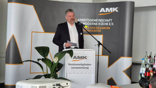 Mann im Anzug hält eine Rede am Rednerpult bei der AMK-Vereinsmitgliederversammlung, neben Mikrofon, Zimmerpflanze und Getränkeflaschen im Konferenzraum