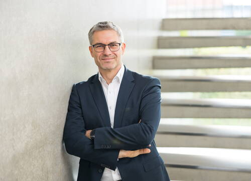 Geschäftsmann mit Brille lehnt lächelnd mit verschränkten Armen an einer Betonwand vor einer modernen Treppe
