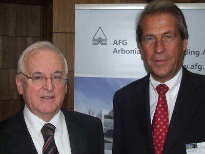 Zwei ältere Geschäftsmänner im Anzug stehen nebeneinander vor einem Firmenbanner in einem Konferenzraum und blicken direkt in die Kamera.