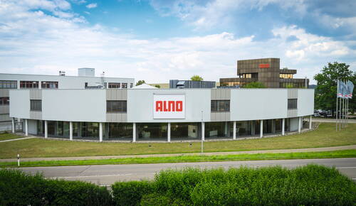 Modernes ALNO-Firmengebäude mit großem Logo an der Fassade, Glasfront im Erdgeschoss und begrüntem Außengelände an einer Straße unter blauem Himmel
