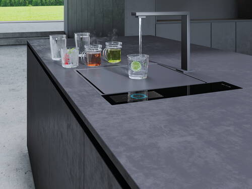 Moderne Kücheninsel mit integrierter Armatur, aus der Wasser in ein Glas mit Limettenscheibe läuft, umgeben von mehreren dampfenden und farbigen Getränken in Glasbechern.