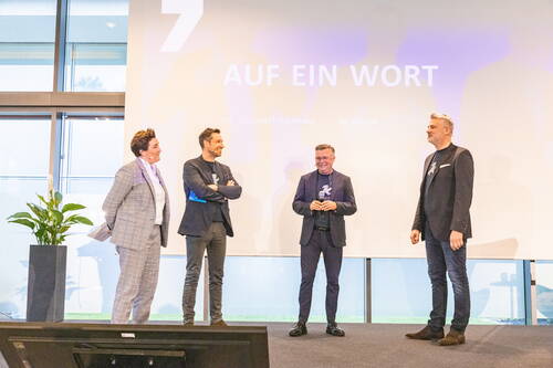 Vier Mitglieder einer Geschäftsführung stehen locker im Dialog auf einer Bühne vor einer Leinwand mit der Aufschrift „Auf ein Wort“ in einem modernen Konferenzraum.
